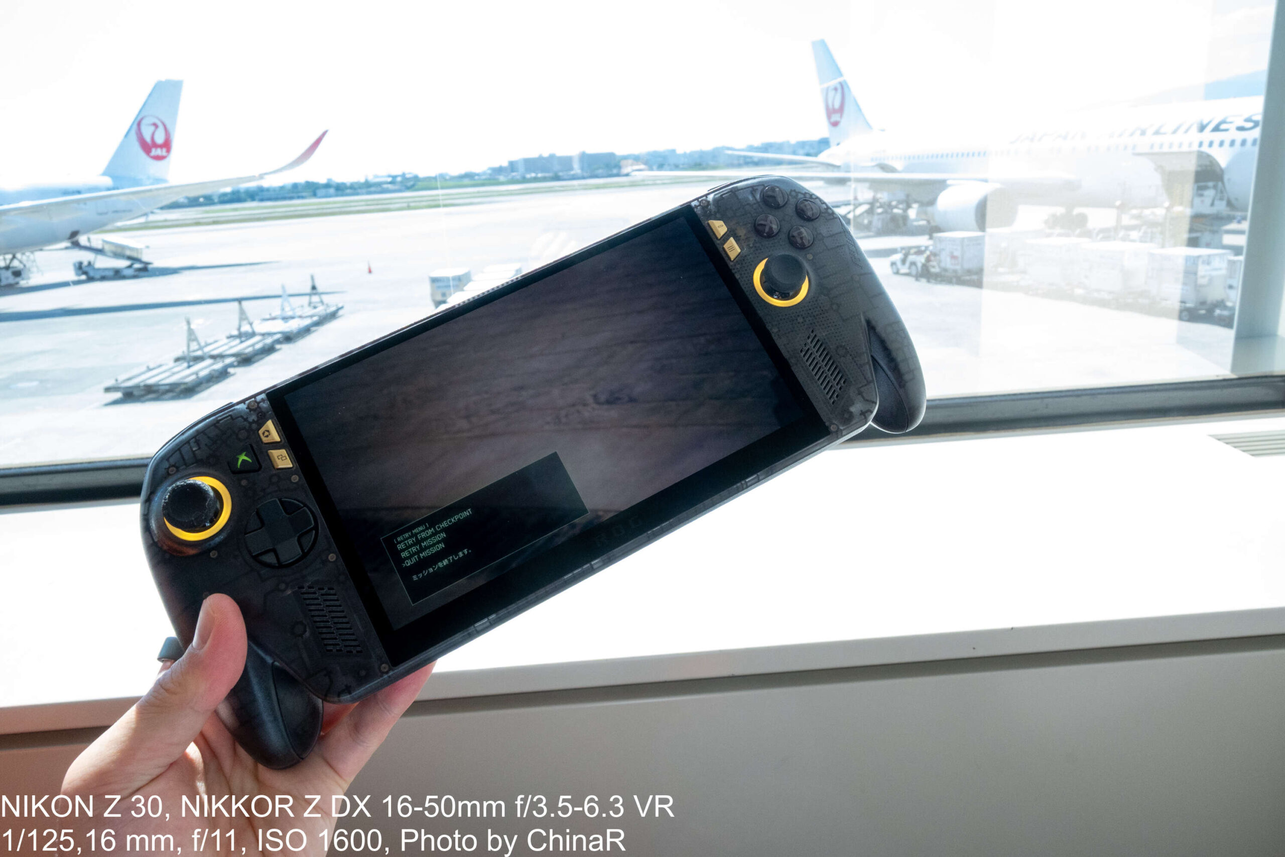 伊丹空港の窓際でROG XBOX Ally X20を手に持つ。窓の外にJALの機体